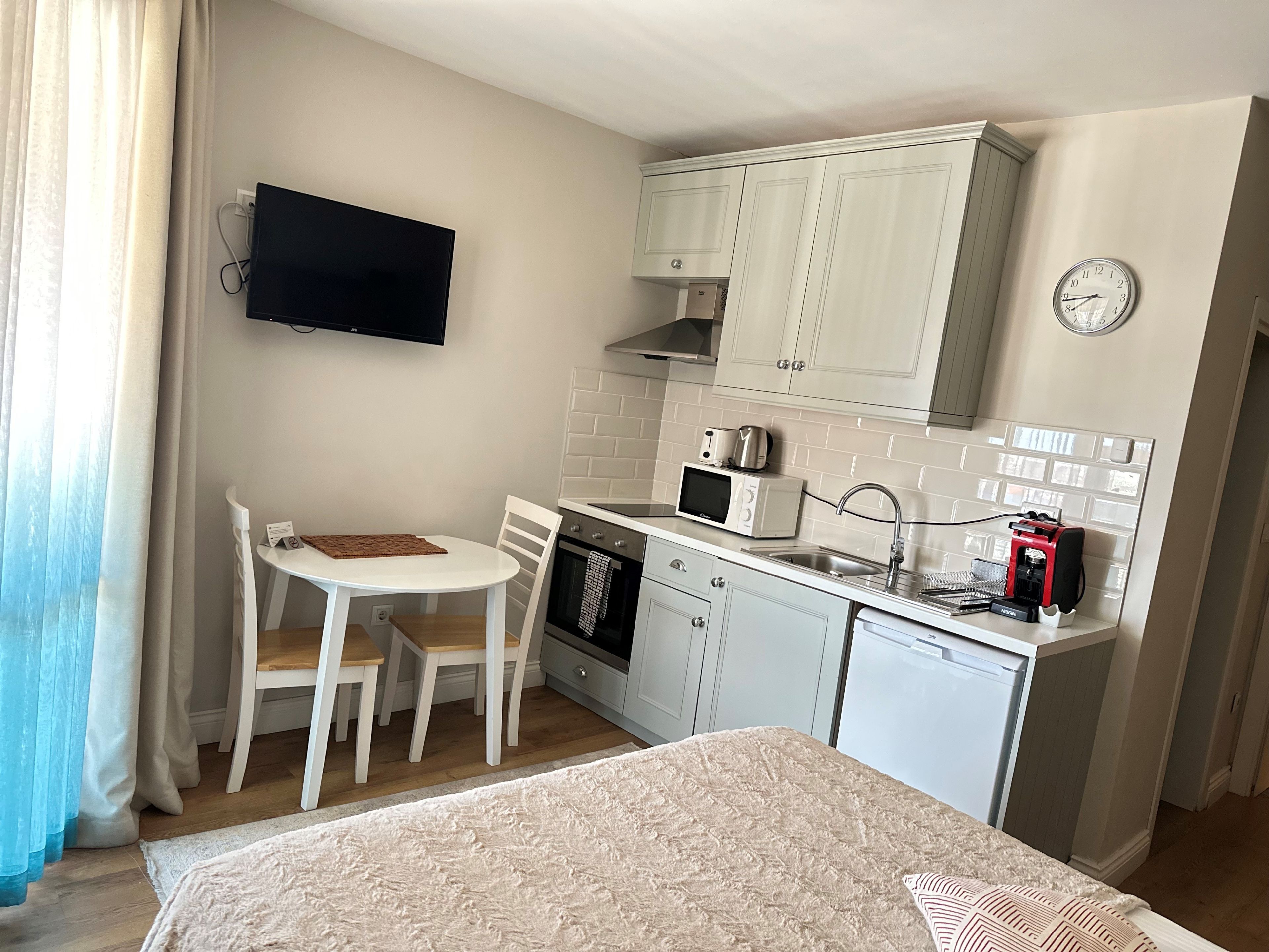 A compact kitchen with white cabinets, a microwave, a coffee machine, and a sink. There is a small dining table with chairs and a TV mounted on the wall.