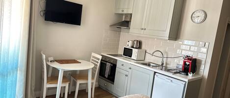 A compact kitchen with white cabinets, a microwave, a coffee machine, and a sink. There is a small dining table with chairs and a TV mounted on the wall.