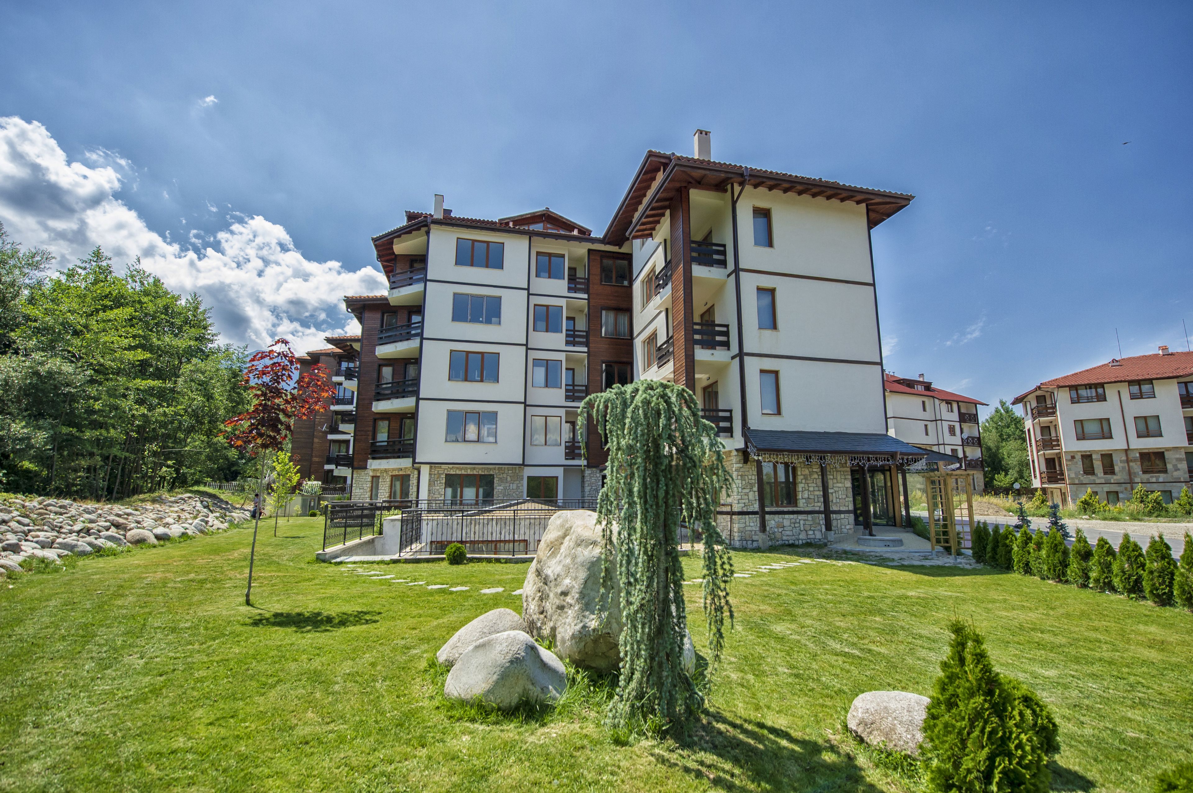 A multi-story residential building with balconies, a stone facade, and a well-maintained lawn with rocks and a weeping willow tree.