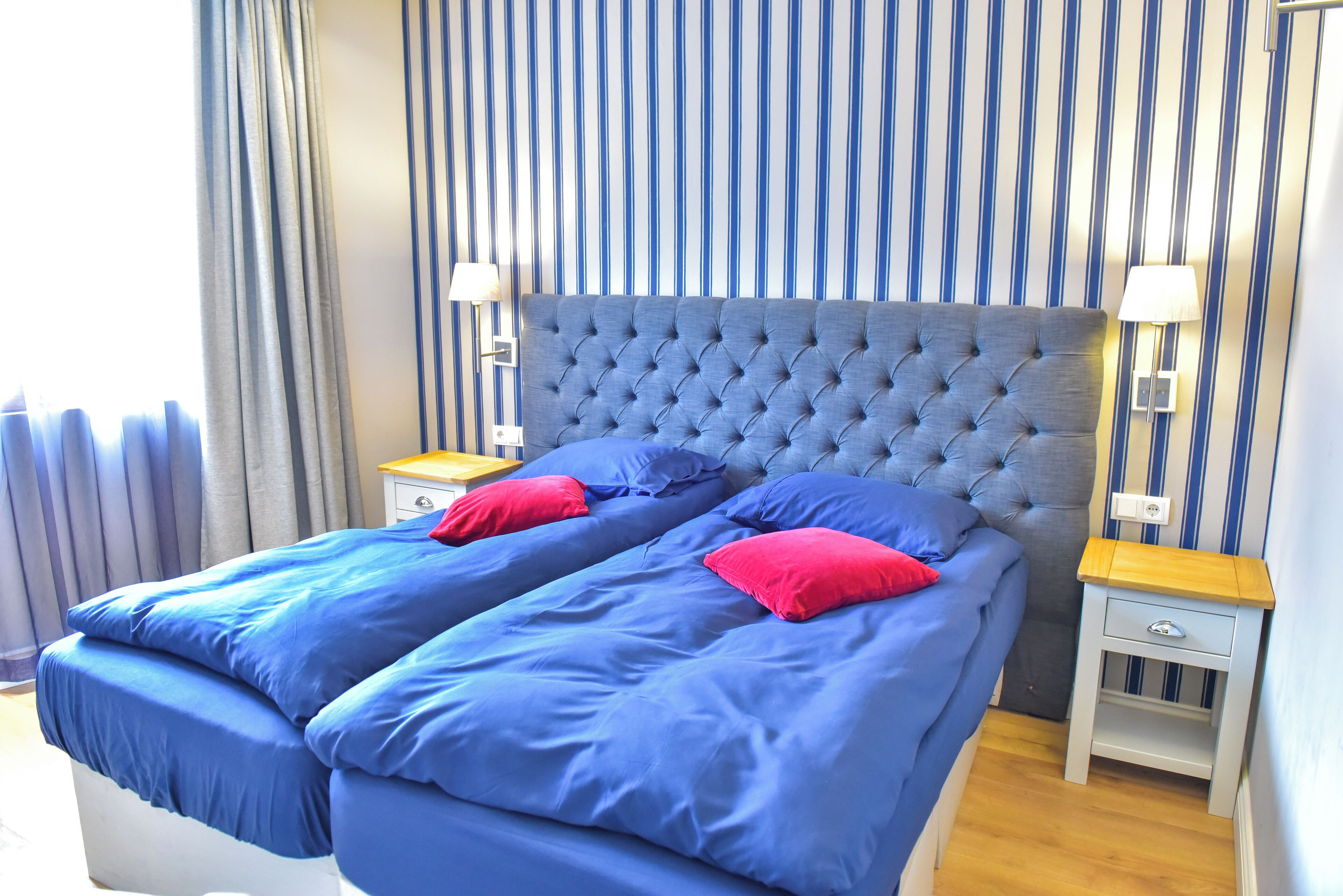 A double bed with blue bedding and red pillows, two bedside tables with lamps, and a striped wall.