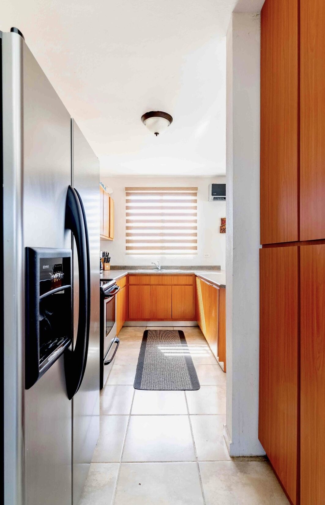 A compact kitchen with a refrigerator, wooden cabinets, a sink, and a window with blinds.