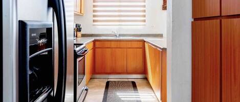 A compact kitchen with a refrigerator, wooden cabinets, a sink, and a window with blinds.