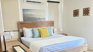 Une chambre moderne avec un lit en bois, une table de chevet, une commode, un climatiseur fixé au mur et un tableau représentant l’océan.