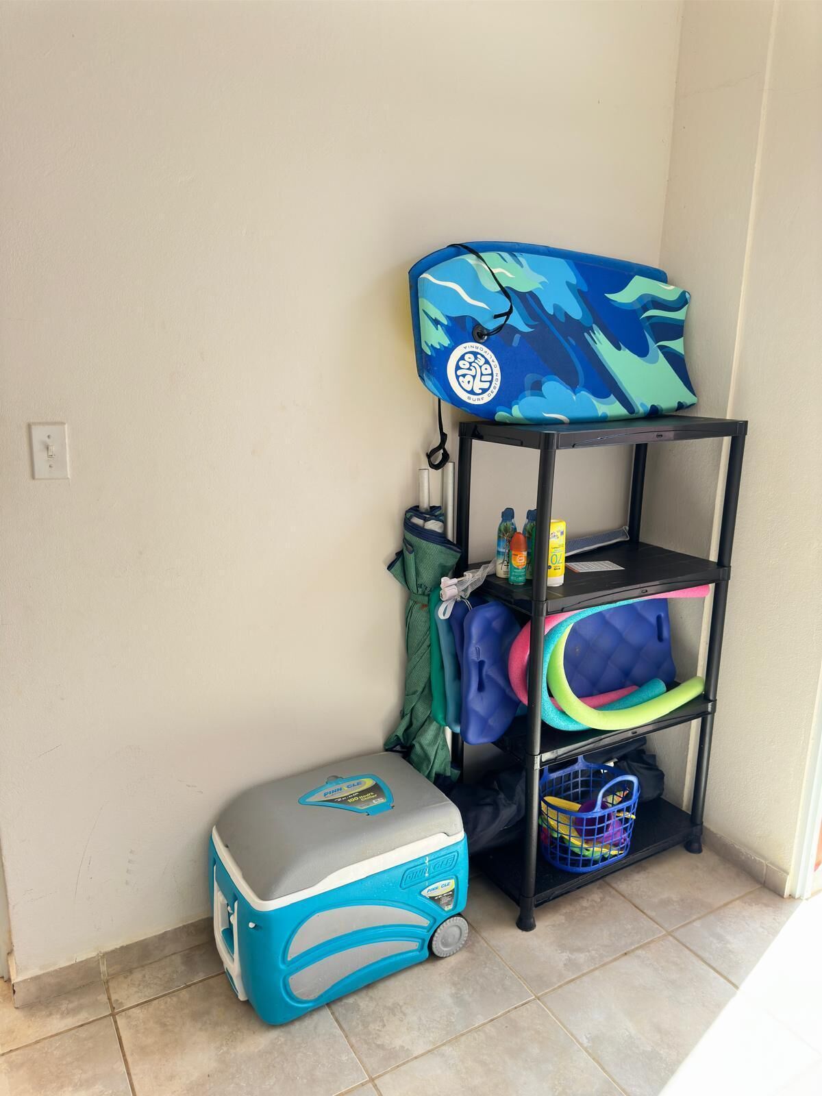 A corner of a room with a blue and white cooler, a black shelf with various items, and a green towel hanging on the wall.