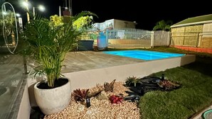 Un espace extérieur aménagé pour la nuit, comprenant une piscine, des plantes en pot et une zone délimitée par une clôture.