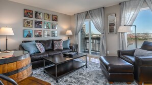 A modern living room with a leather sofa, a coffee table, and a glass-top table with a lamp. There are framed pictures on the wall, a large window with curtains, and a view of the outdoors.