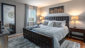 A bedroom with a large bed, a bedside table, a mirror, and a cityscape artwork on the wall.