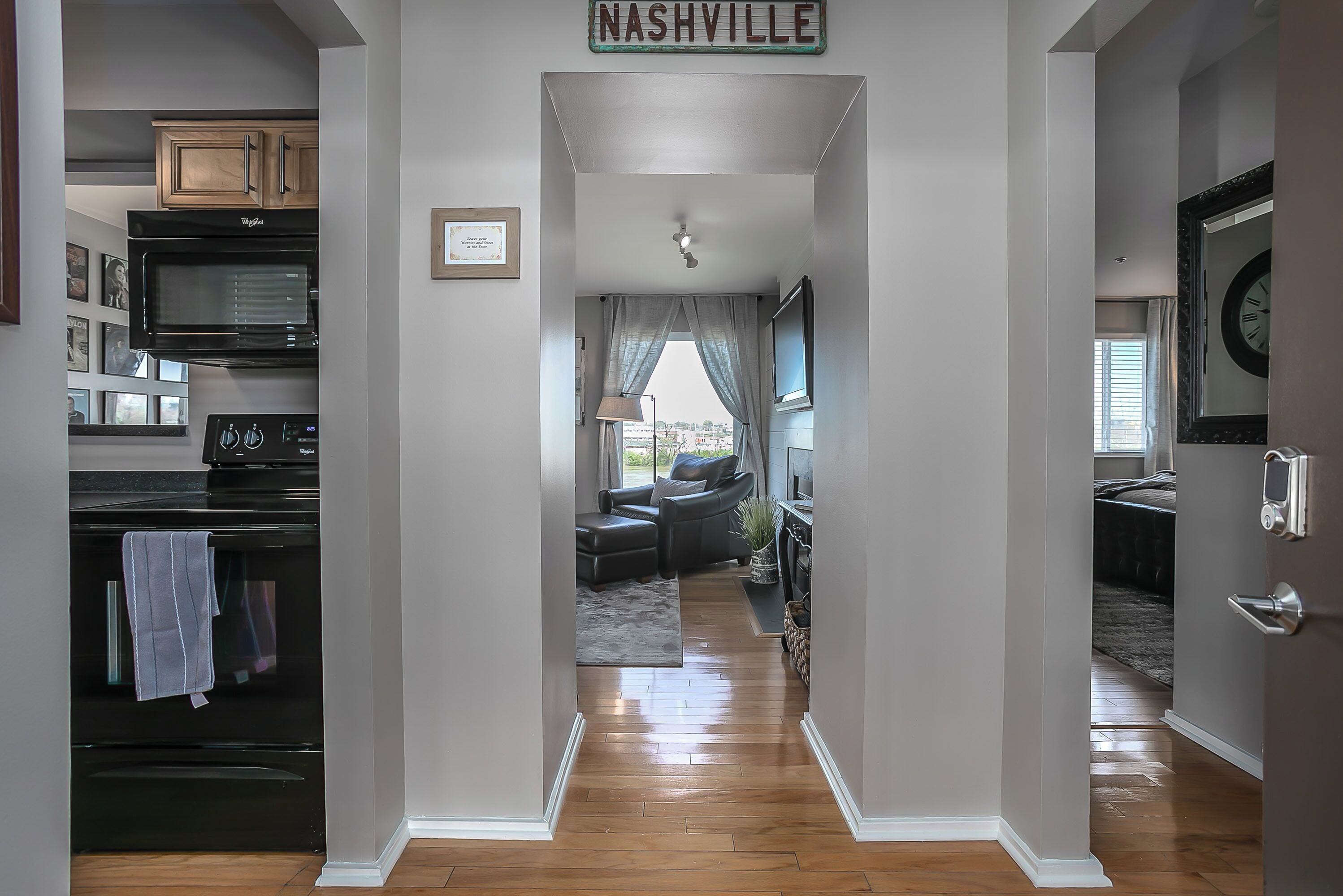 A modern kitchen with a microwave, oven, and wooden cabinets. A hallway leads to a living area with a couch and a view of the city.