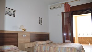 A hotel room with two beds, a wooden headboard, a bedside table with a lamp, and a window with a view of the outside.