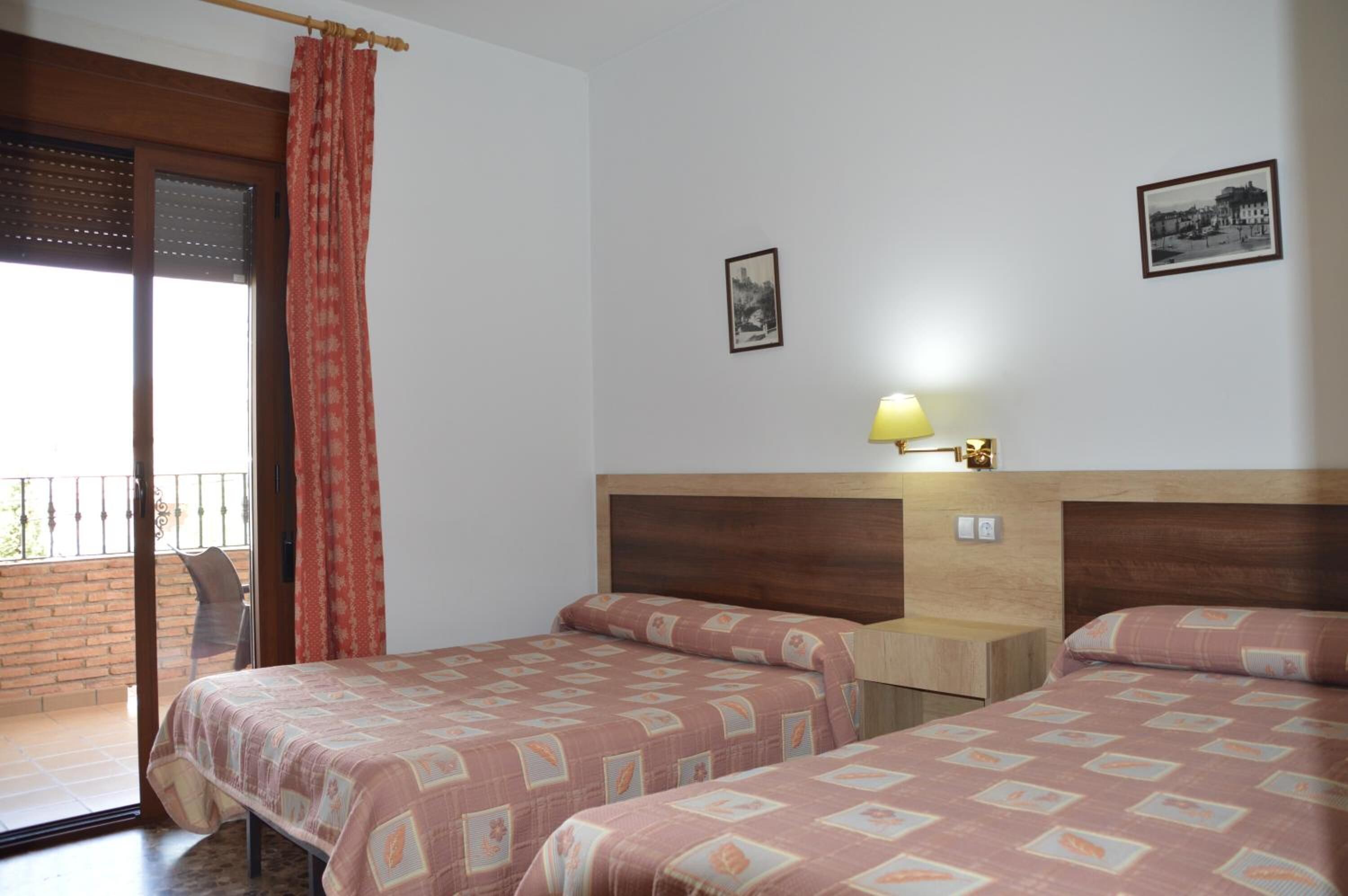 Une chambre d’hôtel avec deux lits, une tête de lit en bois, une petite table de chevet, une lampe et un balcon avec un rideau rouge.