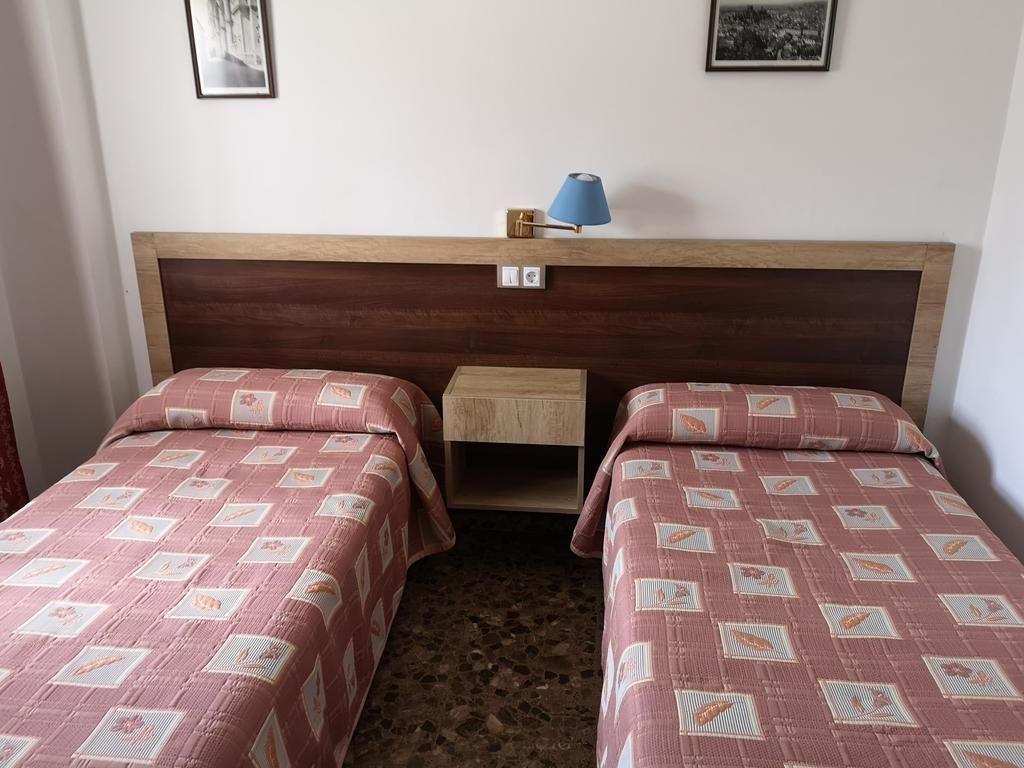 Una habitació amb dos llits individuals, una tauleta de nit, una làmpada i un petit armari de fusta.
