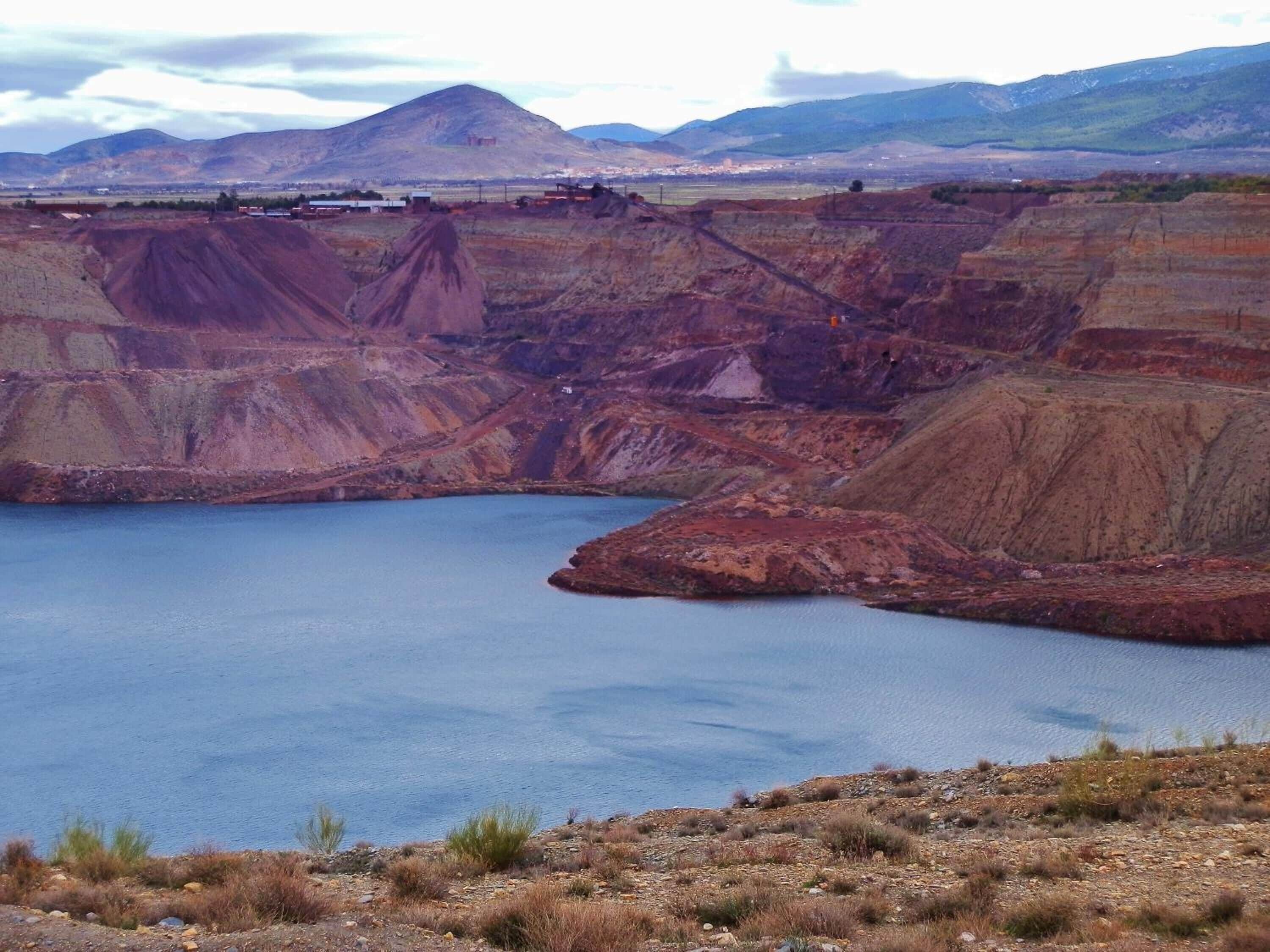 A large open pit with a body of water in the foreground and a mountain in the background.