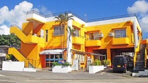 A multi-story building with a bright yellow facade, a visible entrance with a canopy, and a staircase leading to the upper levels. - Pension Orange Box (Miyakojima)