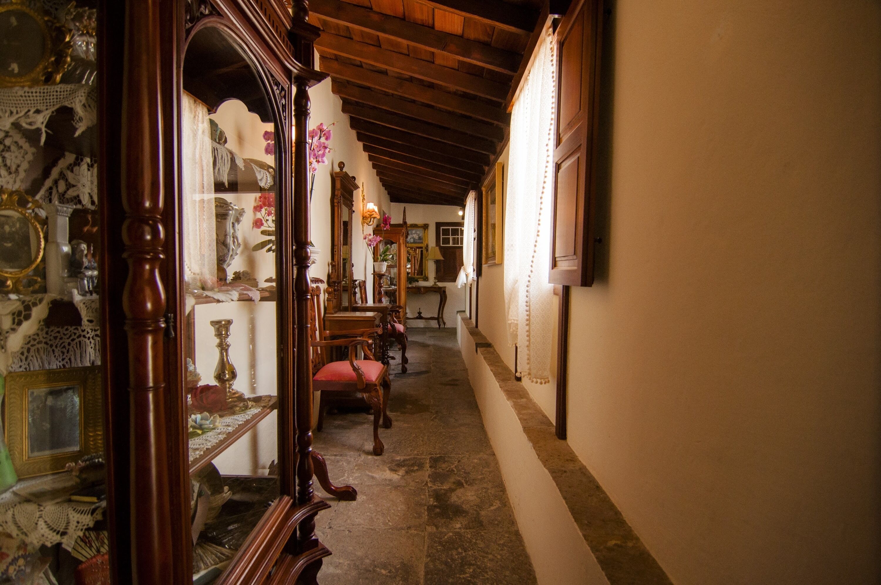 A narrow hallway with wooden flooring and a wooden ceiling. There are several antique chairs and a table with various items on it.