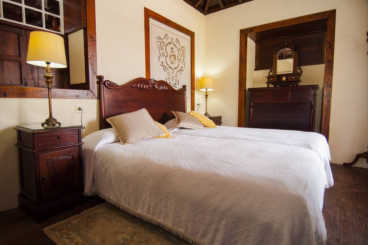 A neatly made bed with a wooden headboard, a nightstand with a lamp, a dresser with a mirror, and a framed artwork on the wall.