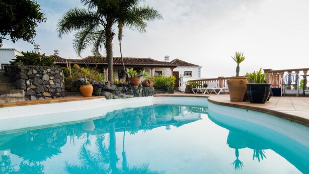 A swimming pool with a stone wall and plants, surrounded by a patio area with lounge chairs. - Finca Arminda (Villa de Mazo)