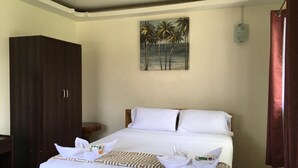 A neatly made bed with white linens, a tropical beach painting above it, and a dark wooden wardrobe to the left. - Aloha Seaside  Resort (San Juan)