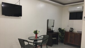 A small, modern room with a TV mounted on the wall, a round dining table with chairs, a small potted plant, a mirror, and a kitchenette area with cabinets and a coffee maker. - Aloha Seaside  Resort (San Juan)