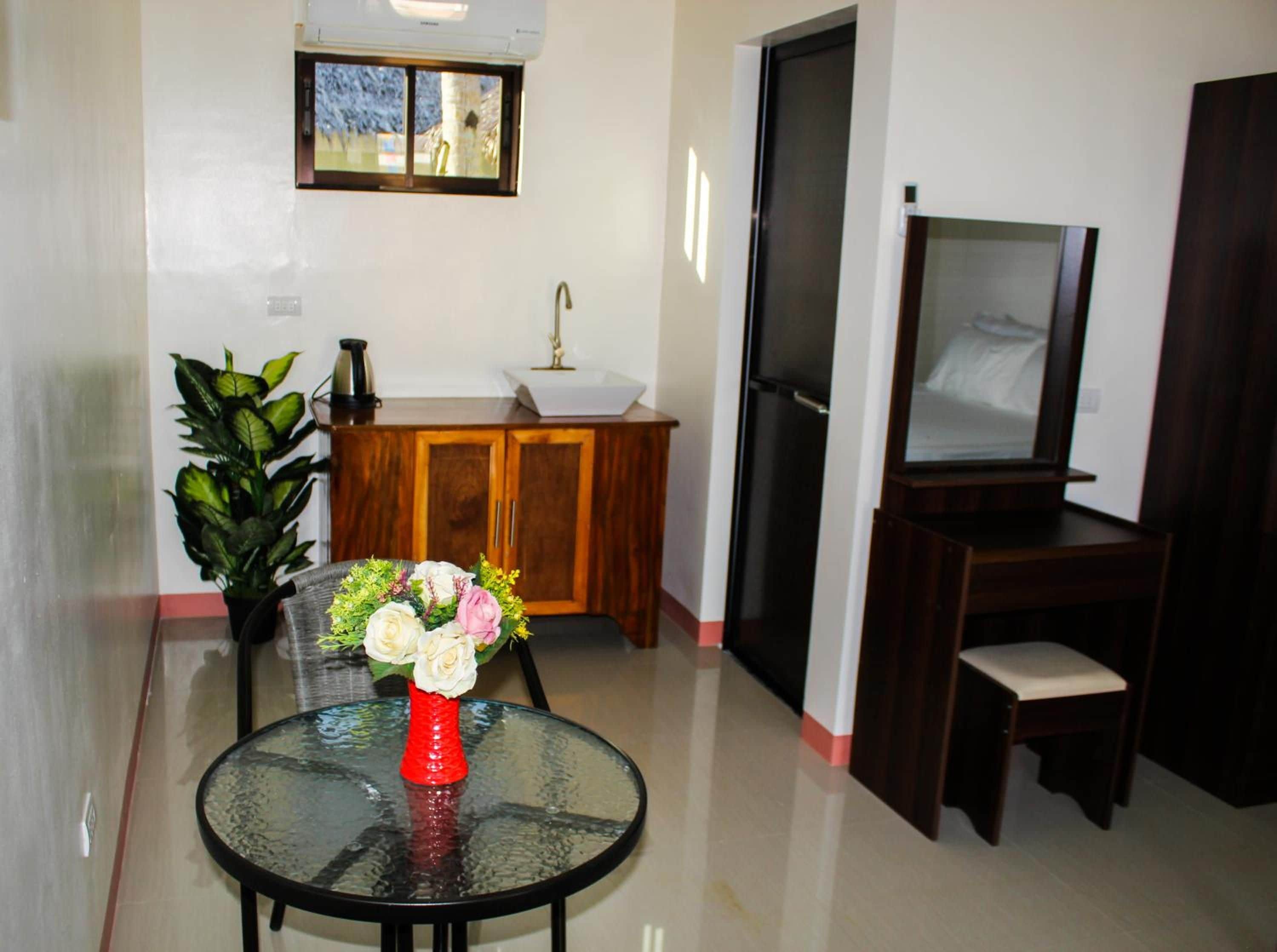 Una habitación con una mesa de vidrio, un jarrón rojo con flores, un armario de madera, un televisor y una planta.