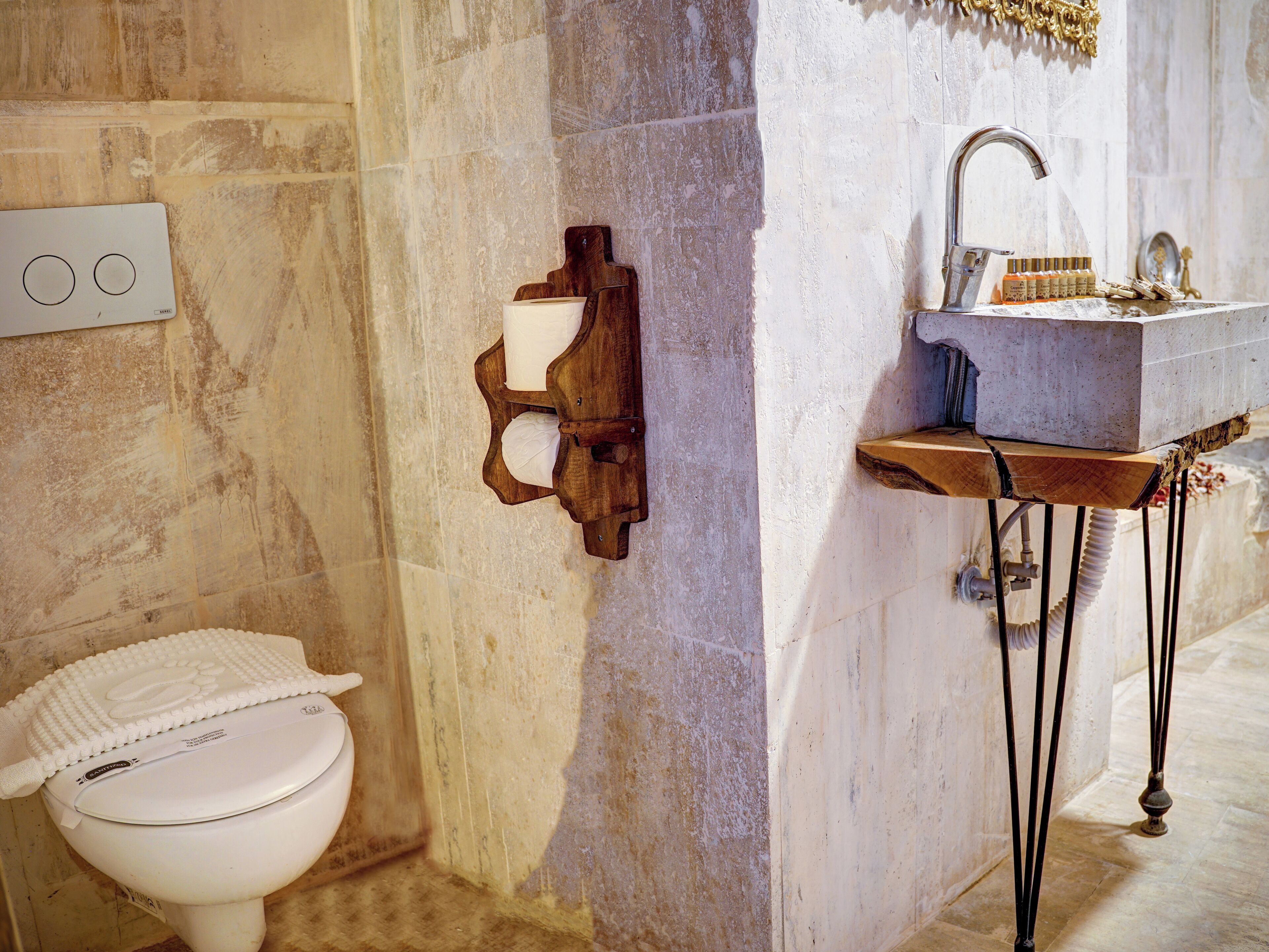 A bathroom with a toilet, a wooden wall-mounted toilet paper holder, a sink with a marble countertop, and a shower area.