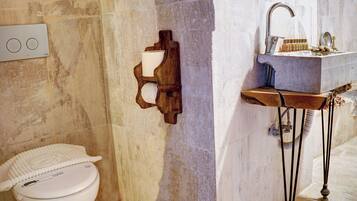 A bathroom with a toilet, a wooden wall-mounted toilet paper holder, a sink with a marble countertop, and a shower area.