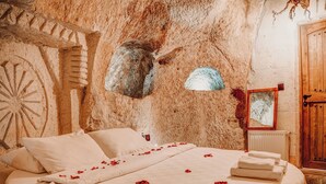 A bed with white linens and red rose petals, a rustic stone wall, and a wooden door. - Mahperi Cappadocia Cave (Nevsehir)