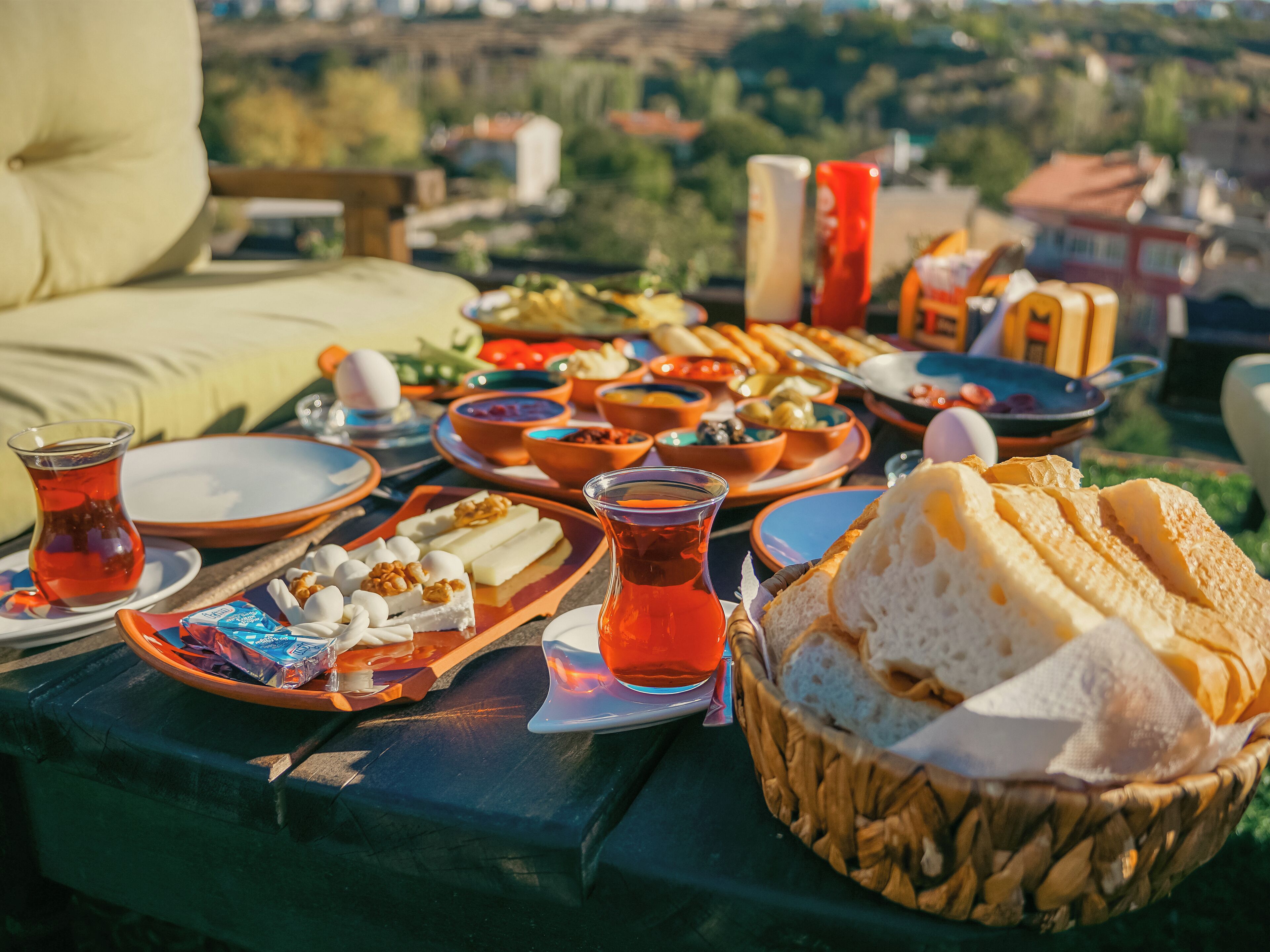 A table set with various dishes, including bread, cheese, and a glass of tea, outdoors with a view of a cityscape.