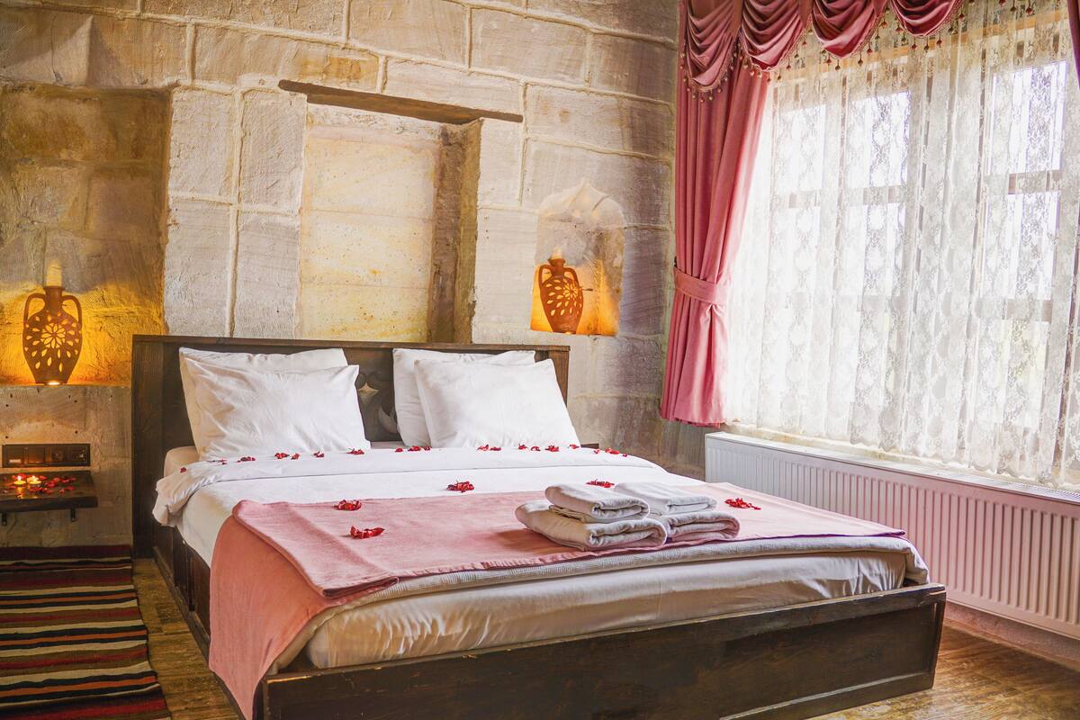 A bedroom with a stone wall, a bed with white linens, a pink curtain, and a wooden floor.