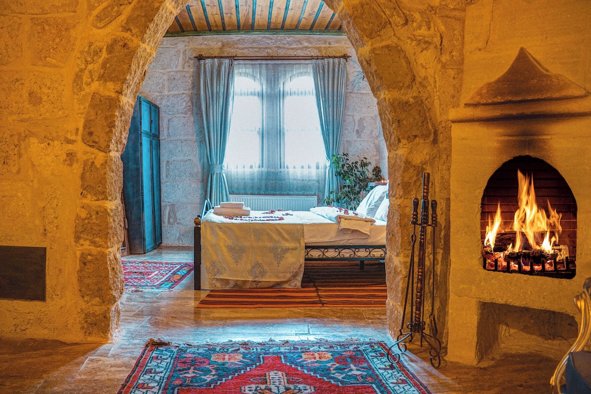 A traditional room with a stone archway, a bed with a patterned bedspread, a fireplace, and a view through the window.