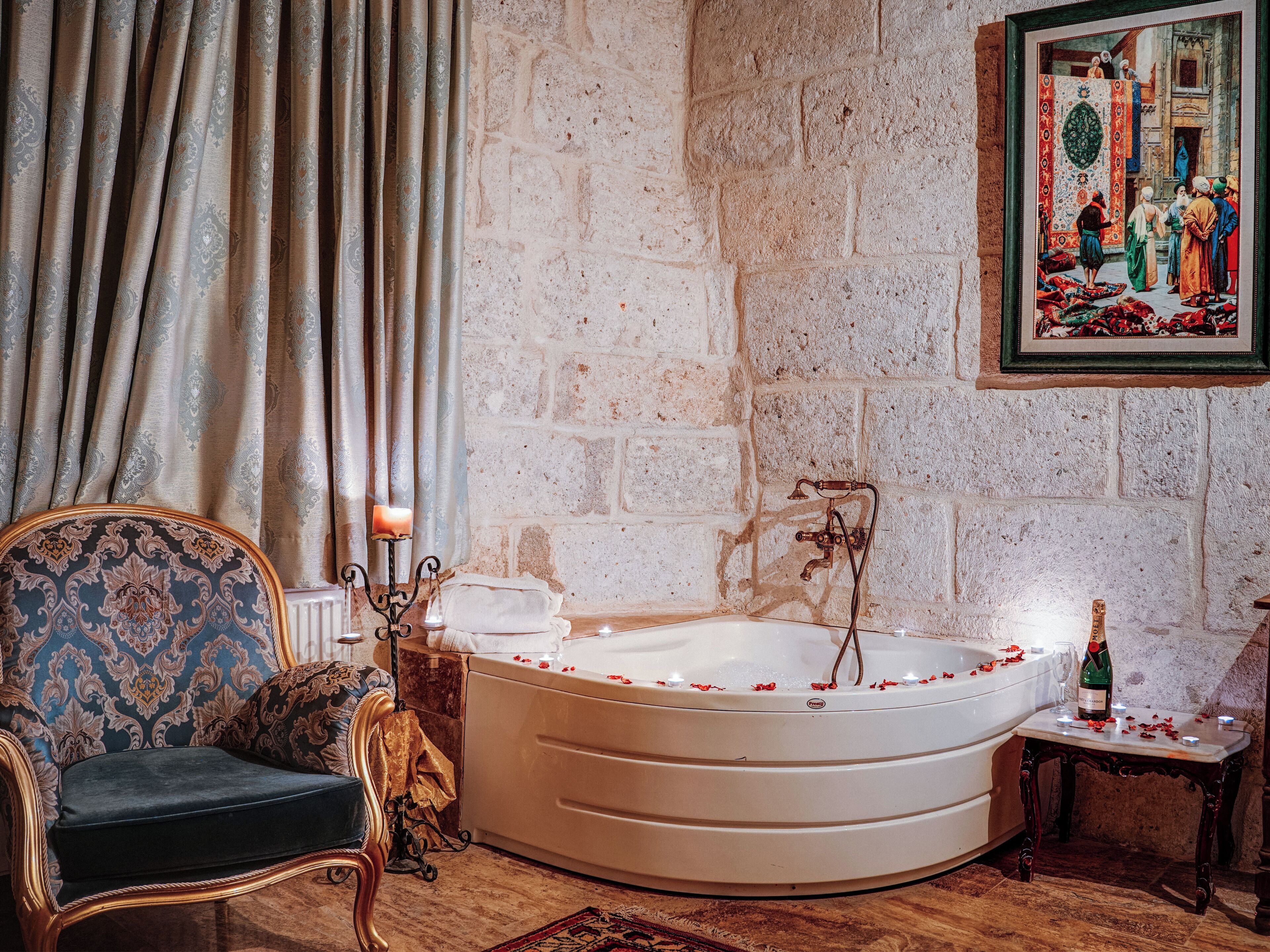 Une pièce avec une grande baignoire, une chaise, une table avec une bouteille et un tableau accroché au mur.