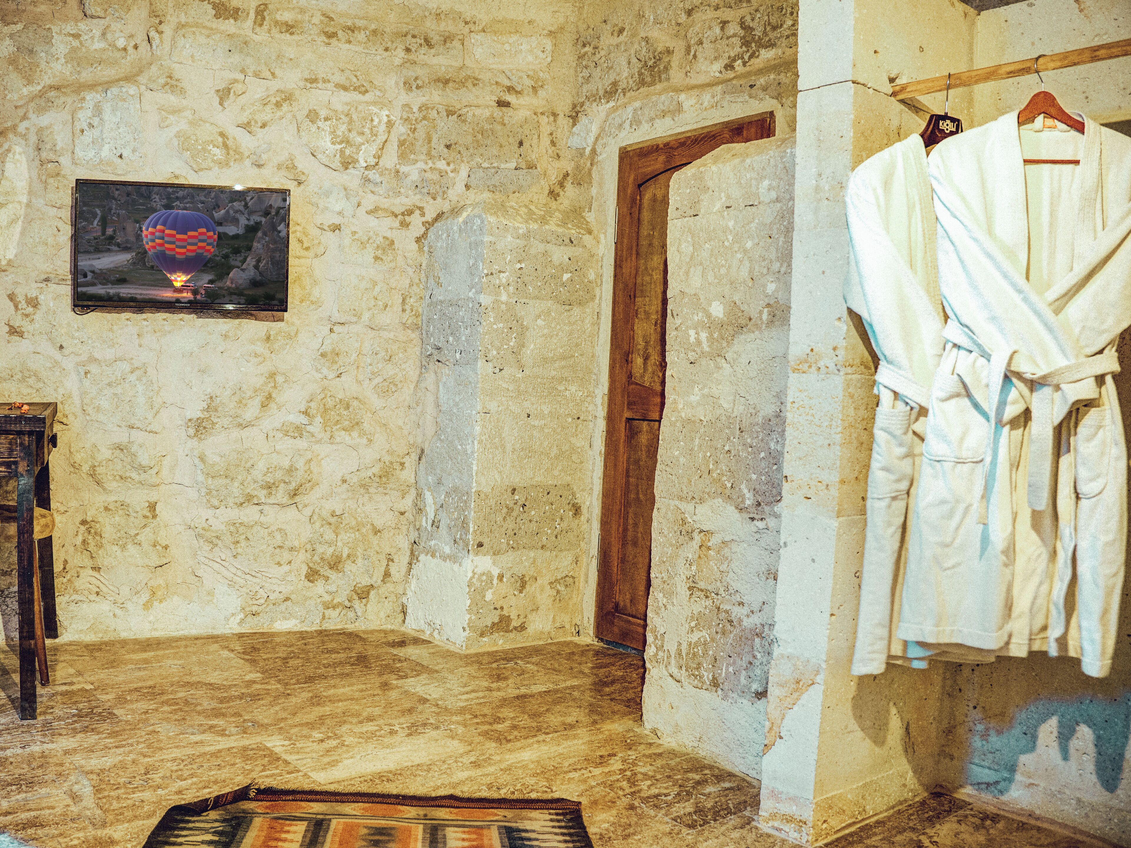 A rustic room with a stone wall, a wooden door, and a framed picture on the wall. There are two white robes hanging on a rack.