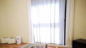 Uma cozinha com bancada de madeira, pia de aço inoxidável, micro-ondas preto e janela com cortinas leves.