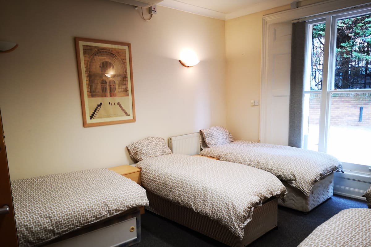 A room with two single beds, a framed picture on the wall, a light fixture, and a window with a view of trees.