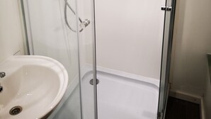 Uma casa de banho com chuveiro em cabine de vidro, uma pia branca e uma porta que dá para outro quarto.