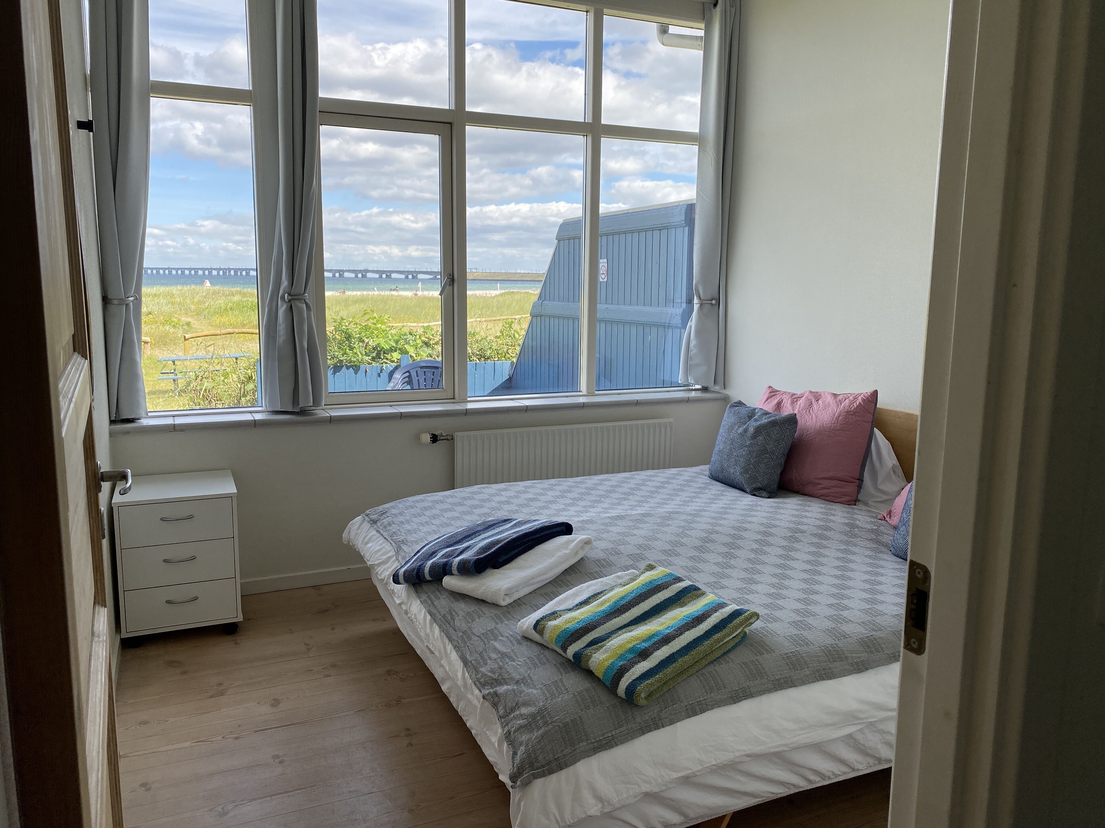 Une chambre avec une grande fenêtre, un lit avec des oreillers, une table de chevet et un radiateur.
