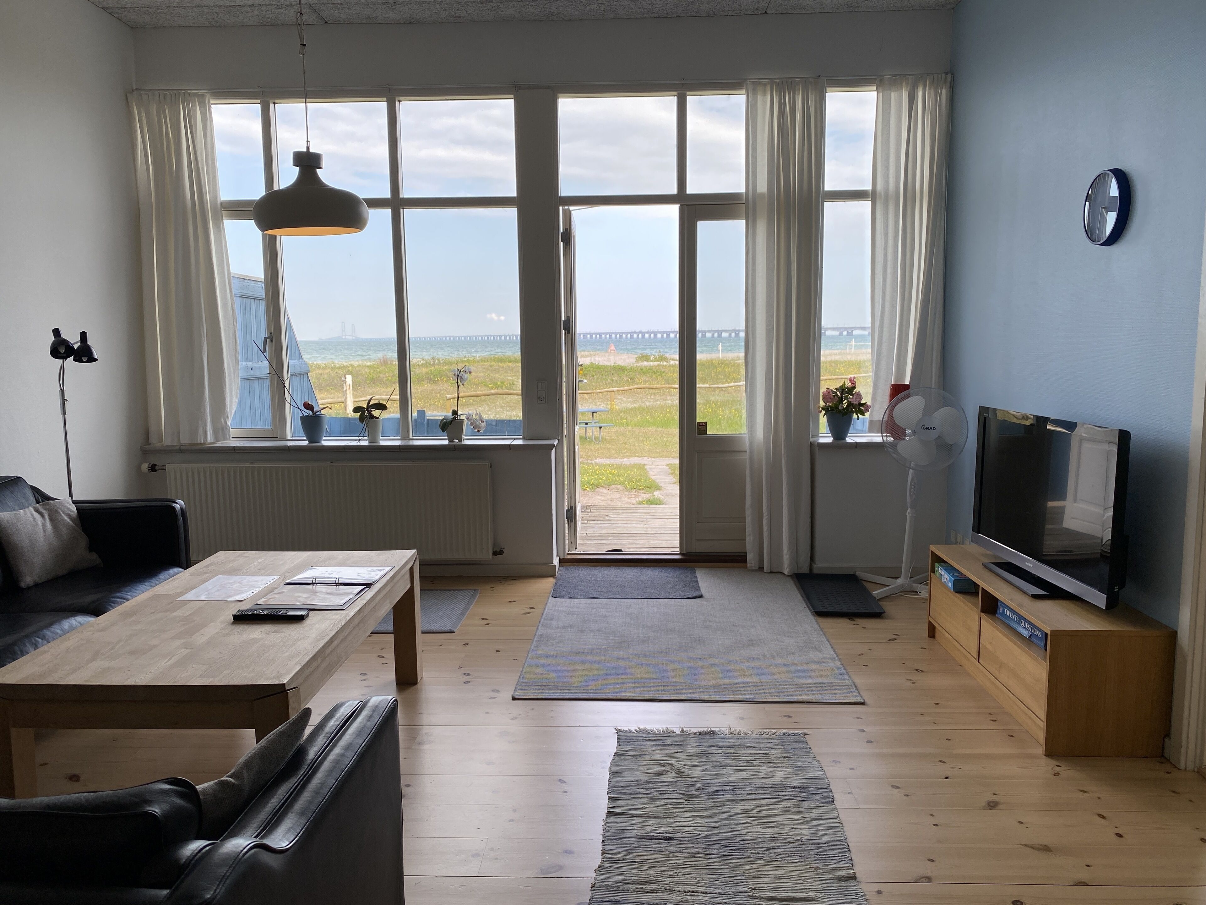 A living room with a sofa, coffee table, TV, and large windows with a view of the beach.