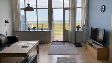 A living room with a sofa, coffee table, TV, and large windows with a view of the beach.
