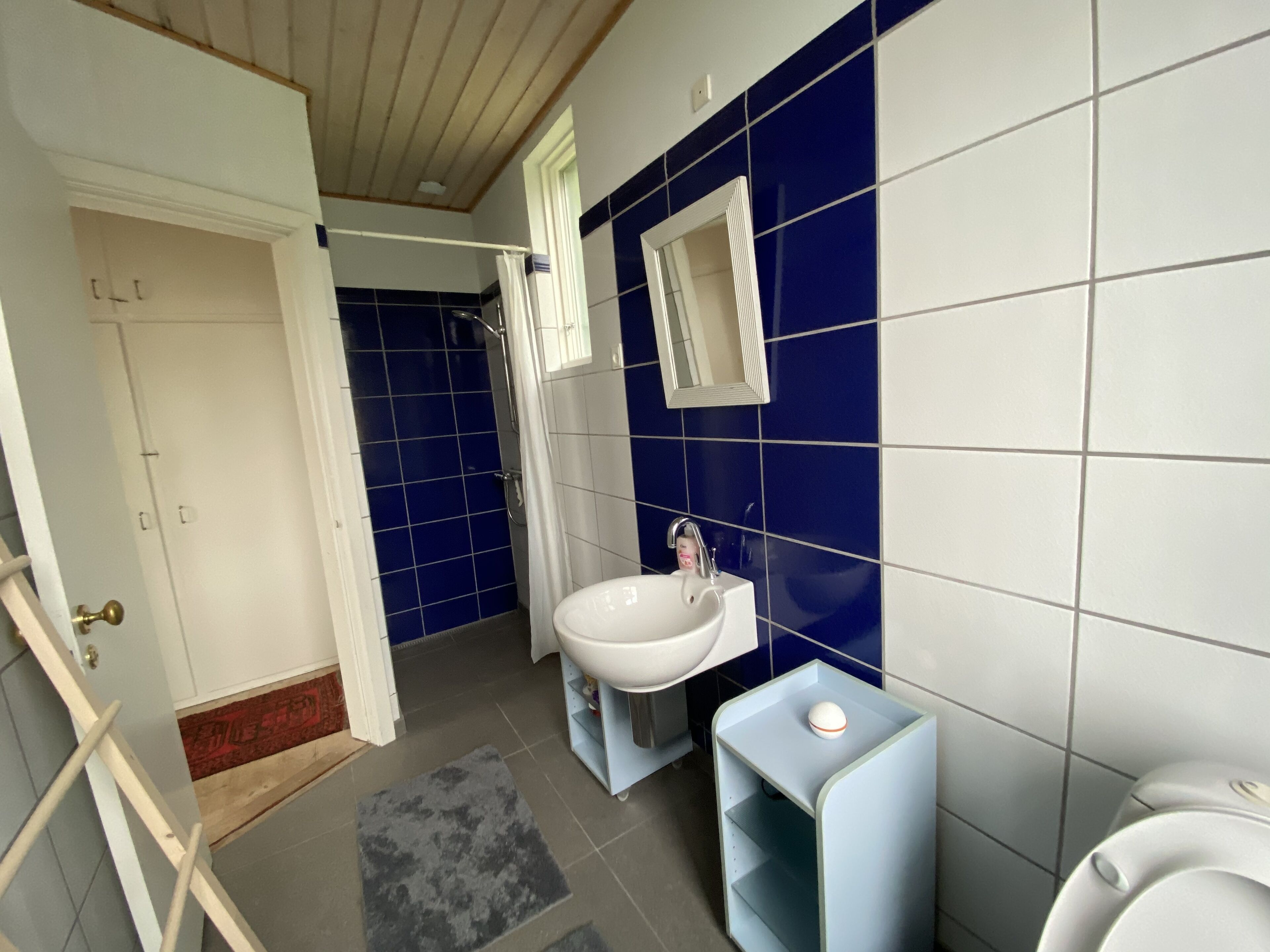 Une salle de bain avec un lavabo blanc, des carreaux bleus et blancs, une petite étagère et une porte donnant sur une autre pièce.