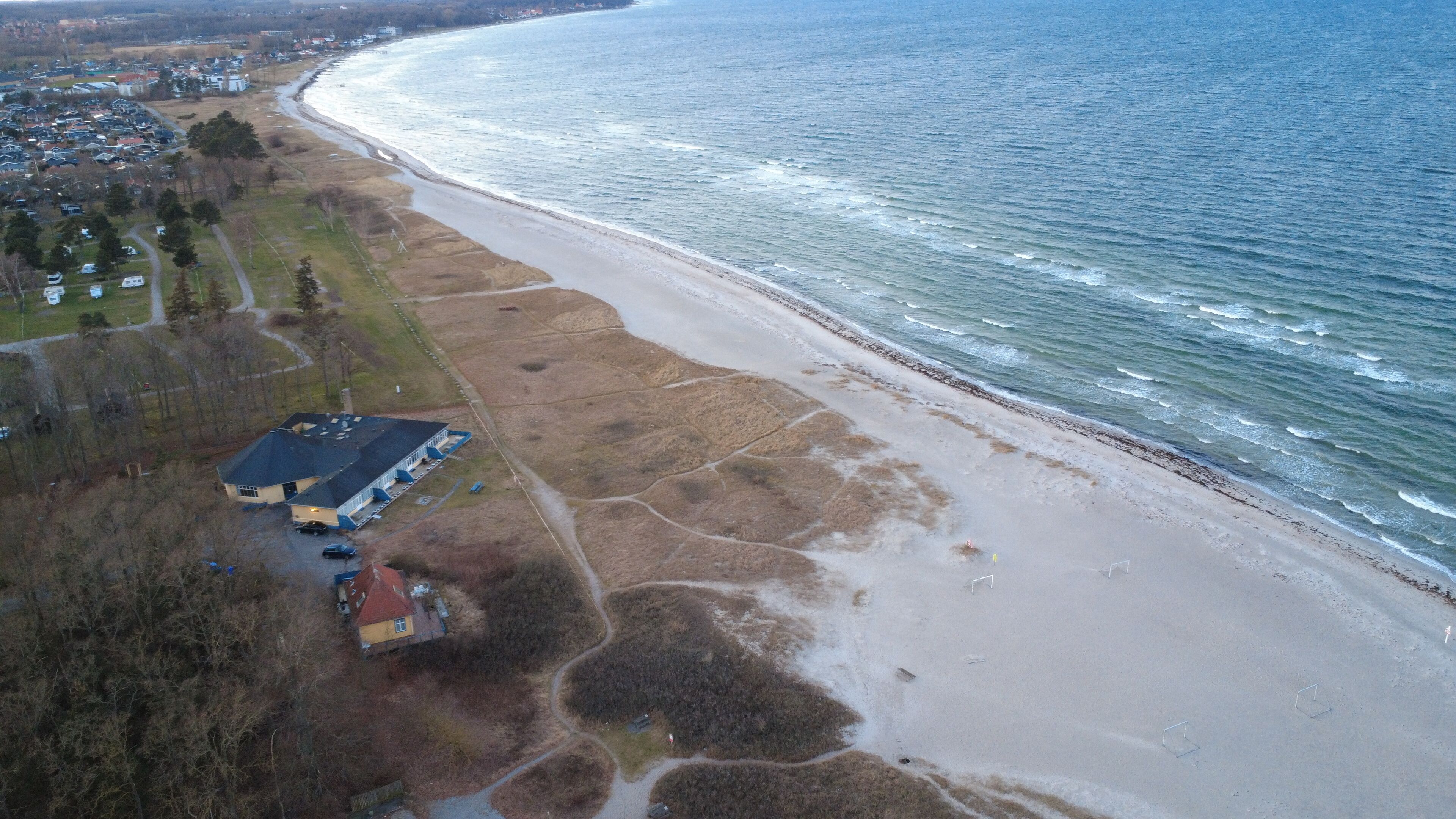 An aerial view of a coastal area with a beach, a residential area, and a building with a blue roof.