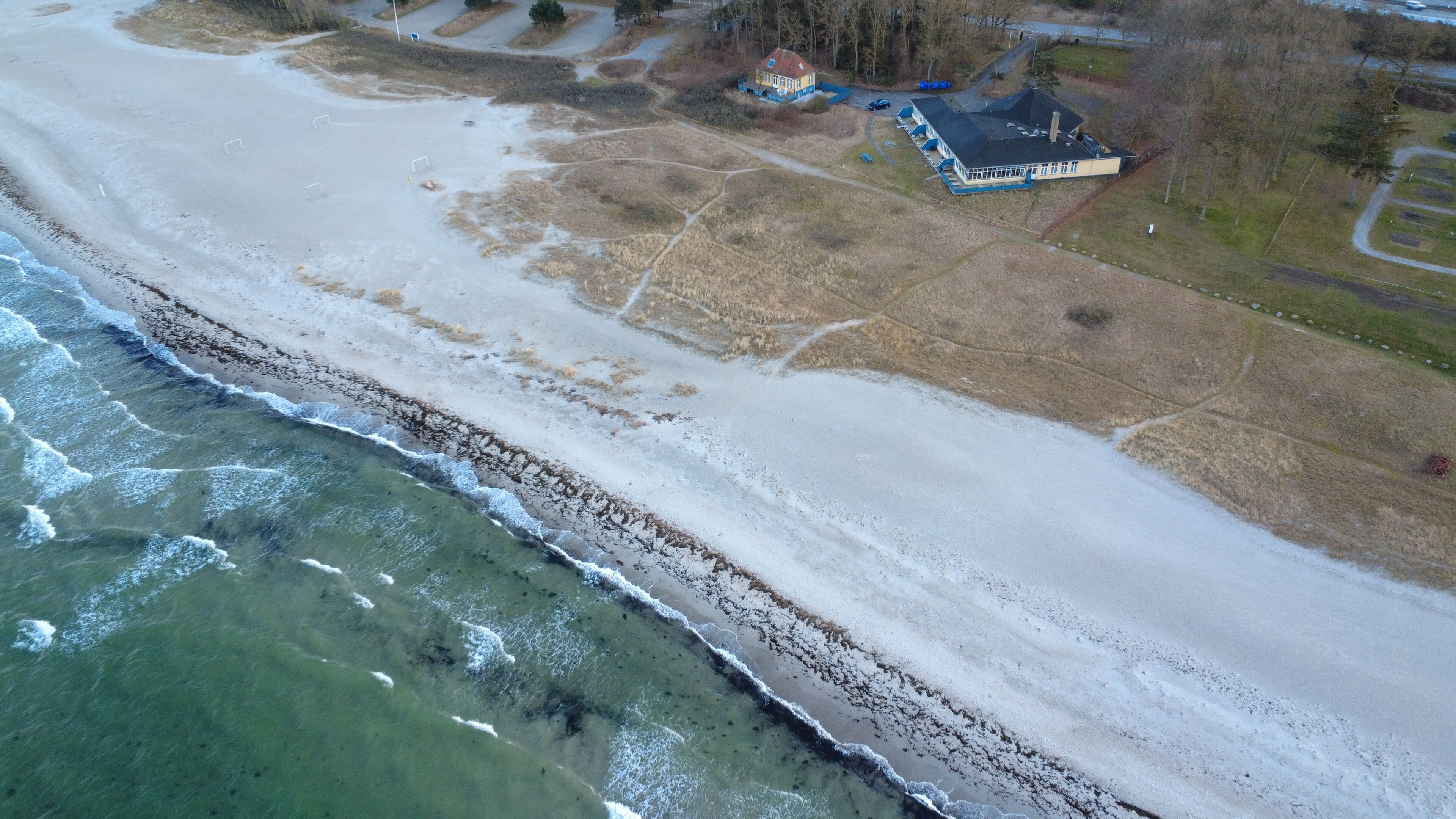 An aerial view of a coastal area with a beach, a building with a blue roof, and a parking lot.