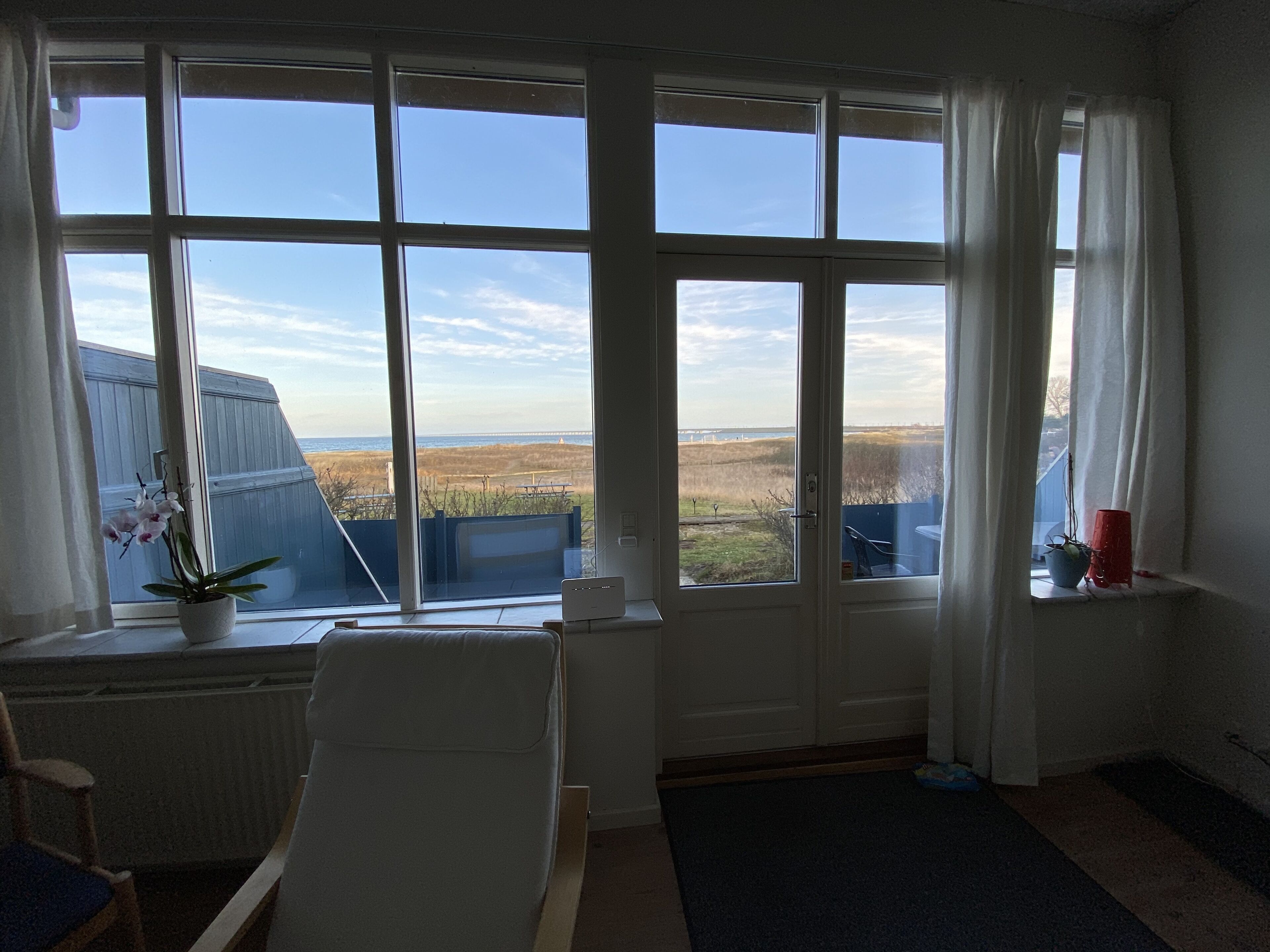 A room with large windows offering a view of the beach and ocean. There is a chair and a small table with a potted plant.