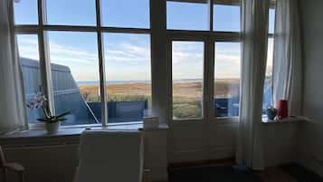 A room with large windows offering a view of the beach and ocean. There is a chair and a small table with a potted plant.