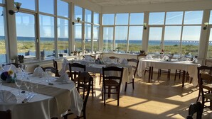 A restaurant with multiple tables set for dining, large windows offering a view of the sea, and a high ceiling. - Skaerven Beachfront Condos and Cottage (Nyborg)