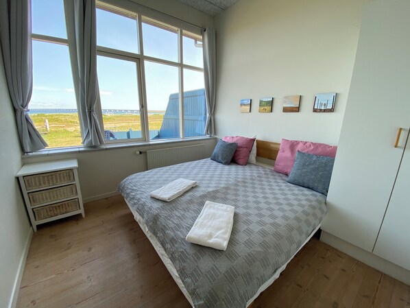 A bedroom with a large bed, a window with curtains, a radiator, and a view of the beach. - Skaerven Beachfront Condos and Cottage (Nyborg)