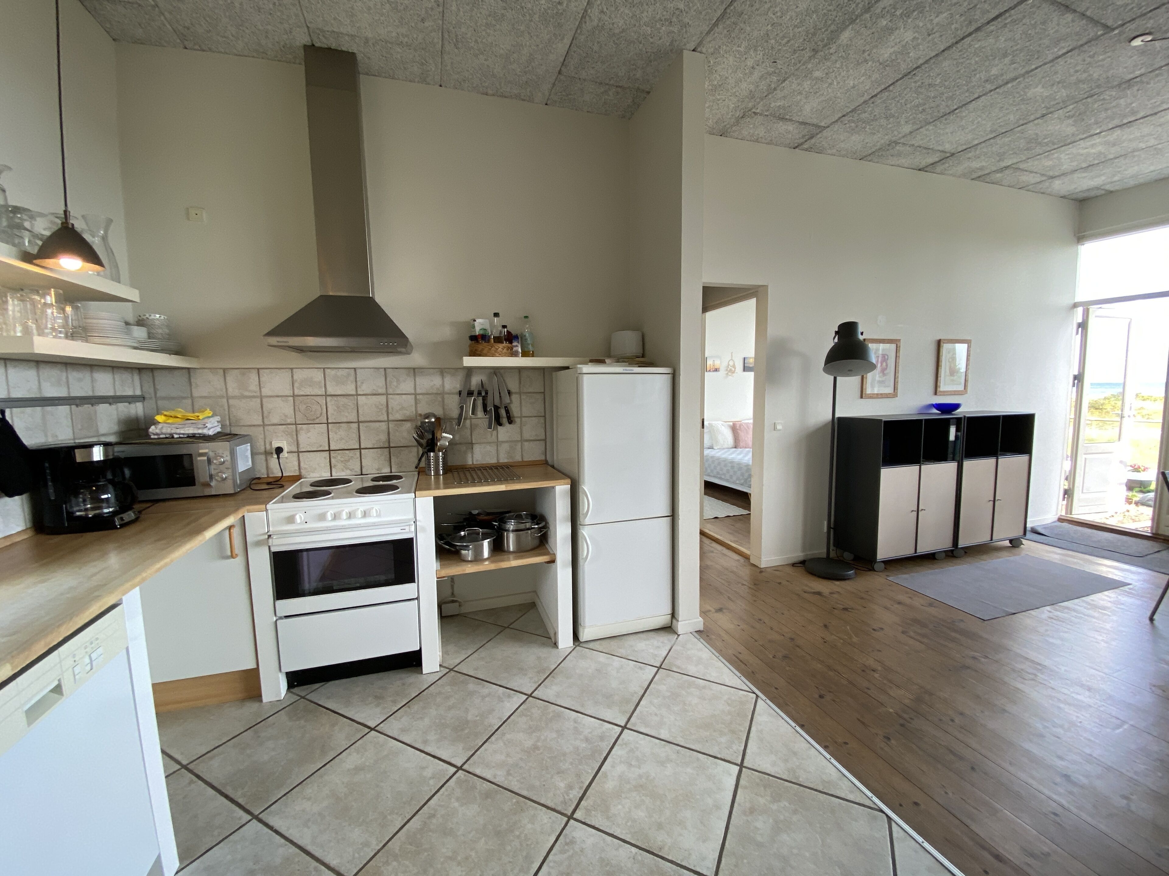 Een moderne keuken met een fornuis, magnetron en koelkast. Er is een houten aanrecht en een betegelde vloer. Een gang leidt naar een slaapkamer met een bed en een deur.