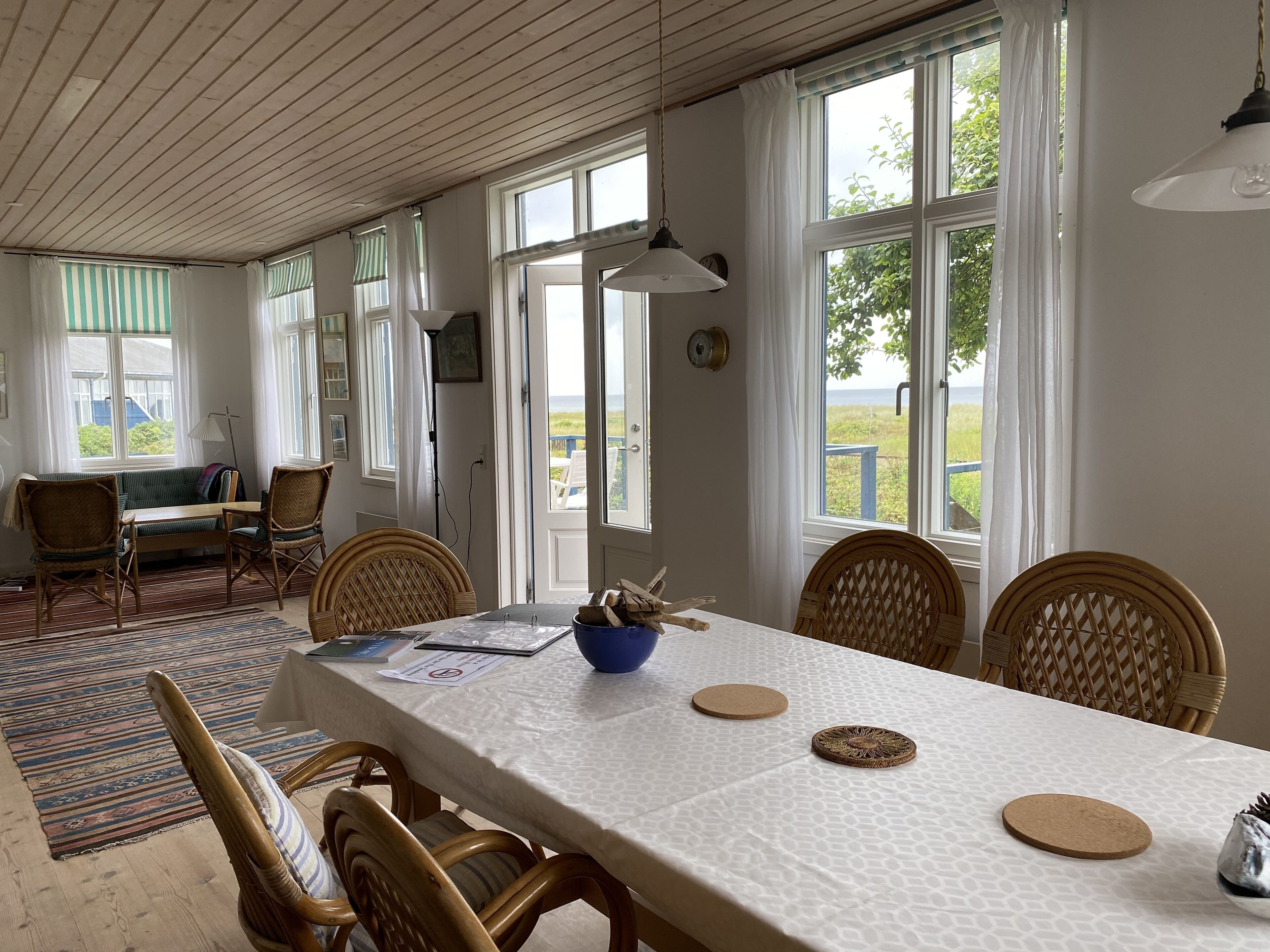 Un comedor con una mesa dispuesta para cuatro personas, rodeada de sillas de mimbre, un tapete estampado y vistas al exterior a través de amplias ventanas.