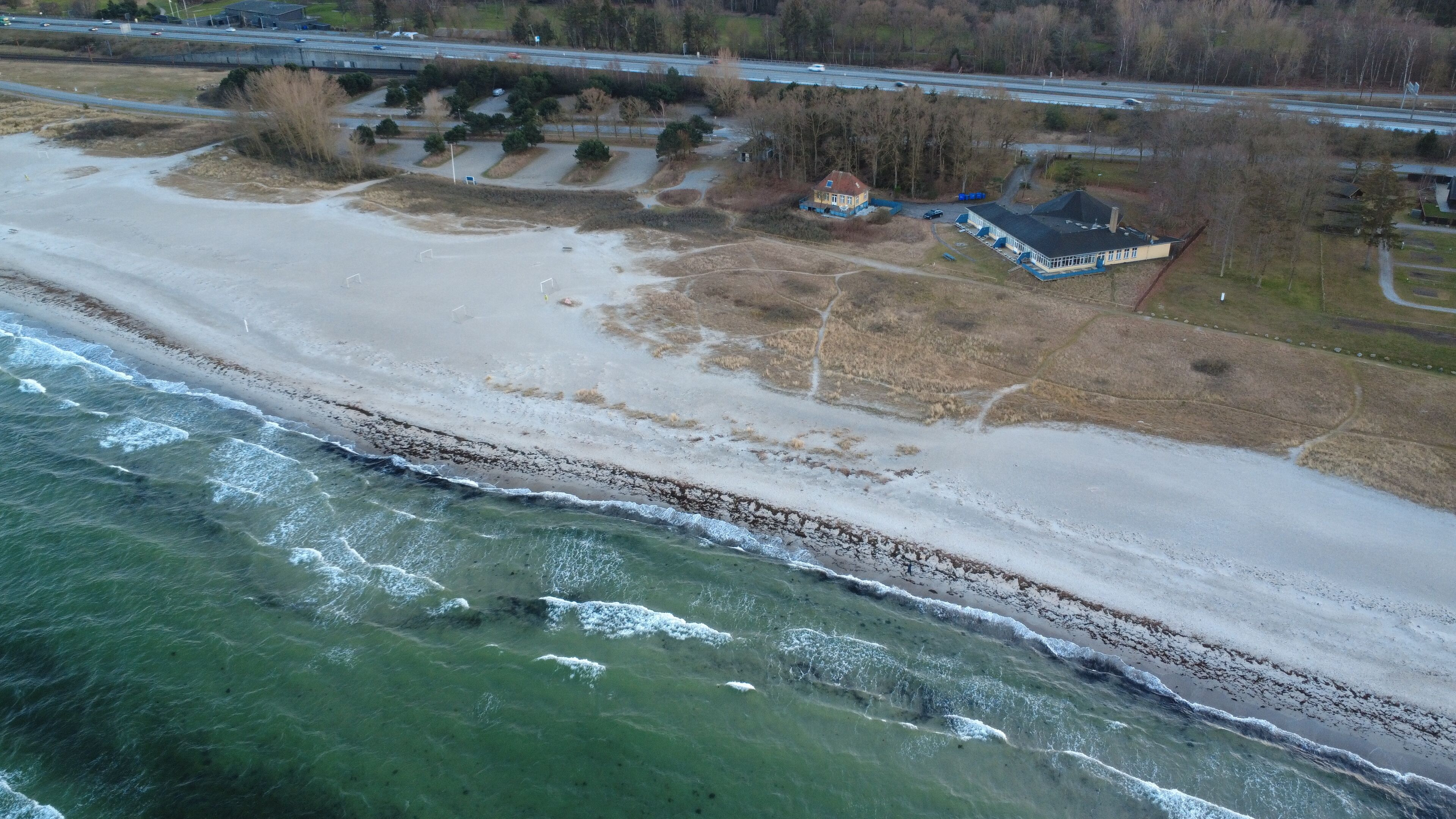 An aerial view of a beach with a house near the shore, a road, and a bridge in the background.