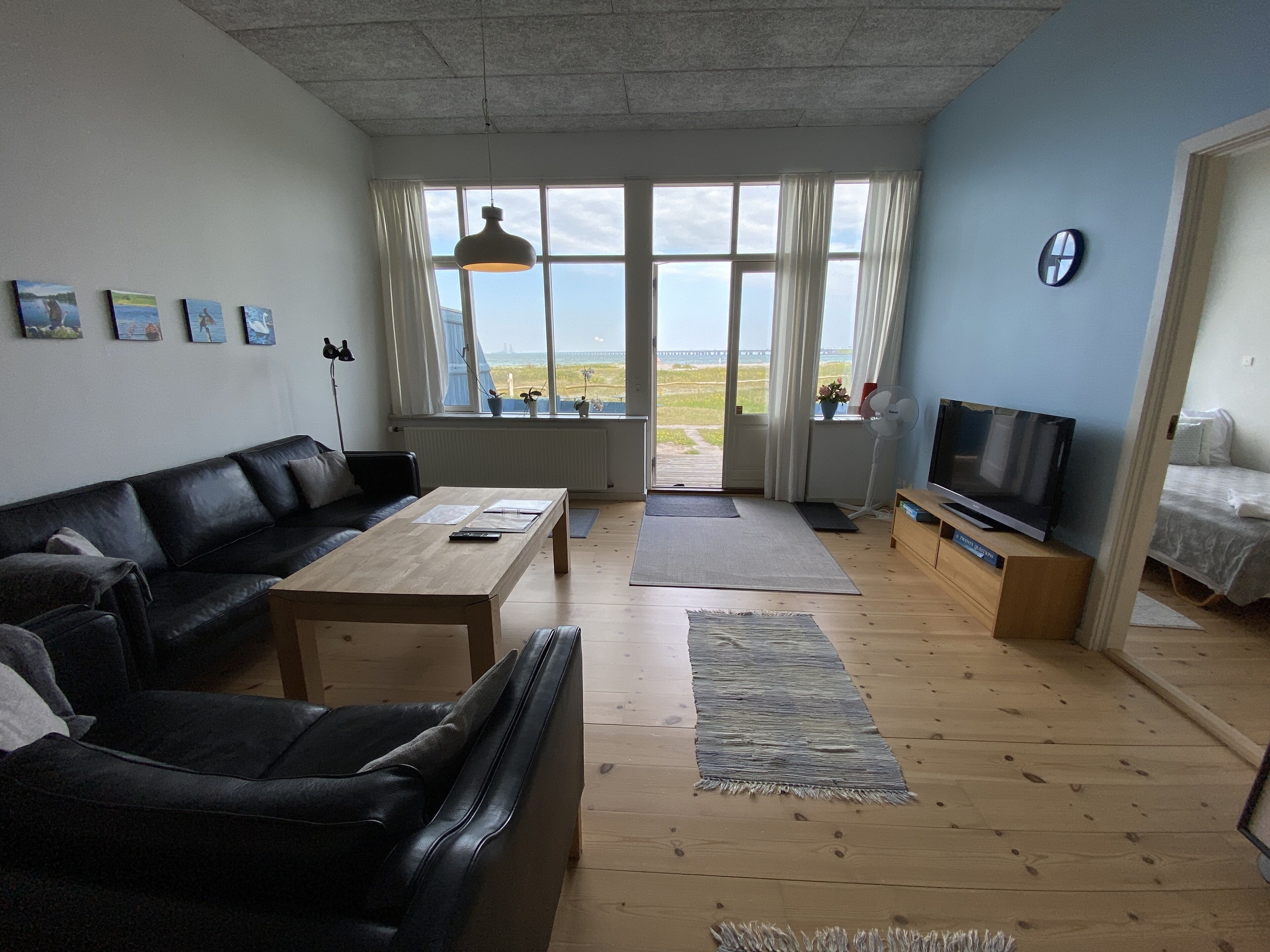 A living room with a black leather couch, a wooden coffee table, a TV on a wooden stand, and a large window with a view of the outdoors.