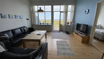 A living room with a black leather couch, a wooden coffee table, a TV on a wooden stand, and a large window with a view of the outdoors.