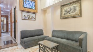 Eine moderne Lobby mit zwei grauen Sofas, einem Glastisch, einer an der Wand montierten Überwachungskamera und einem gerahmten Kunstwerk mit arabischer Kalligraphie.